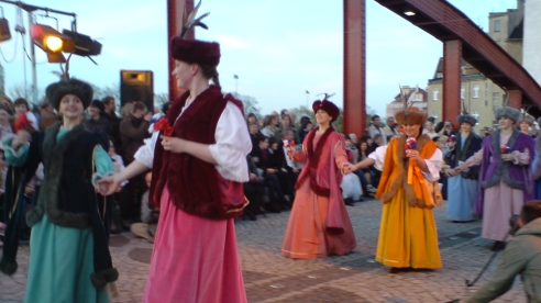 Mostem przechodzą pary tnacerek przebrane w stroje szlachcianek. W tle stoi publiczność.