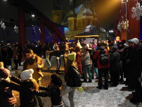 Zgromadzeni na moście ludzie tańczą. W tle widać kościół oraz czerwone przęsła mostu.