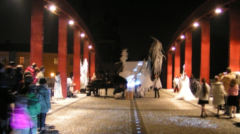 Widok na most. Po obu stornach widać czerwone przęsła i kolumny mostu. Na środku mostu niewyraźny zarys białych postaci z piórami oraz czarny fortepian, przy którym siedzi mężczyzna. Po bokach widać zgoromadzoną publicznosć. Zdjęcie wykonane nocą.