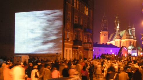 Zdjęcie w nocy. Mało wyraźne. Widać niewyraźny tłum ludzi przechodzący ulicą. W tle widać kamienicę. W głębi widac budynek kościoła.