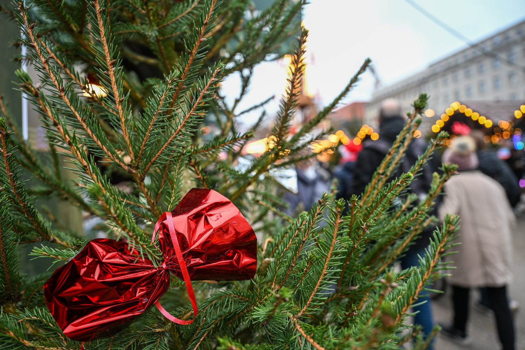 Zbliżenie na gałązkę choinki udekorowaną dużą, błyszczącą ozdobą z czerwonej folii w kształcie zawiniętego cukierka. W rozmytym, nastrojowym tle widać fragmenty jarmarku świątecznego z sylwetkami przechodniów oraz świąteczne iluminacje.