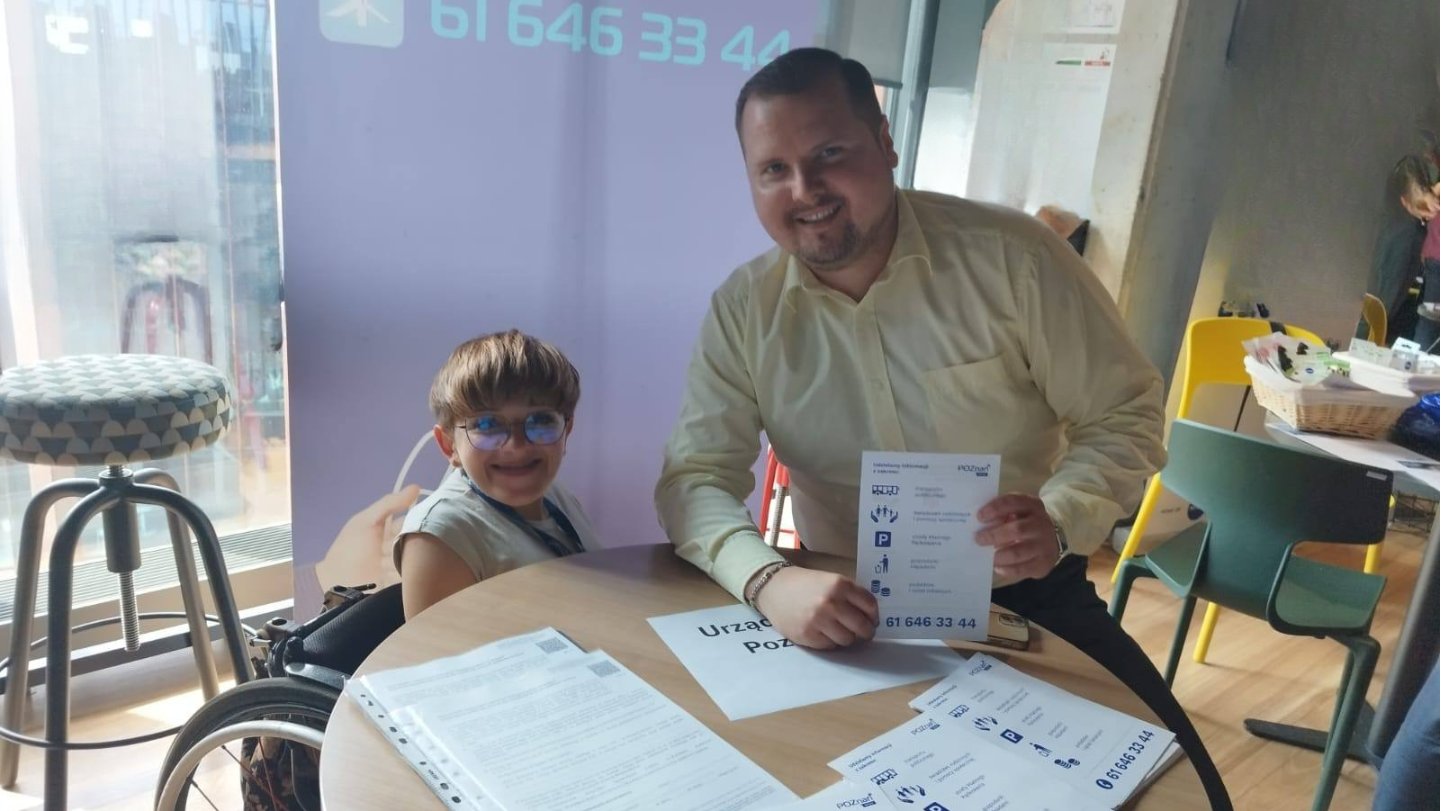 Zdjęcie przedstawia pracowników Biura Poznań Kontakt na Targach Pracy dla Osób z (Nie)pełnosprawnościami, na tle rollupu, z ulotkami na stoliku i w ręce