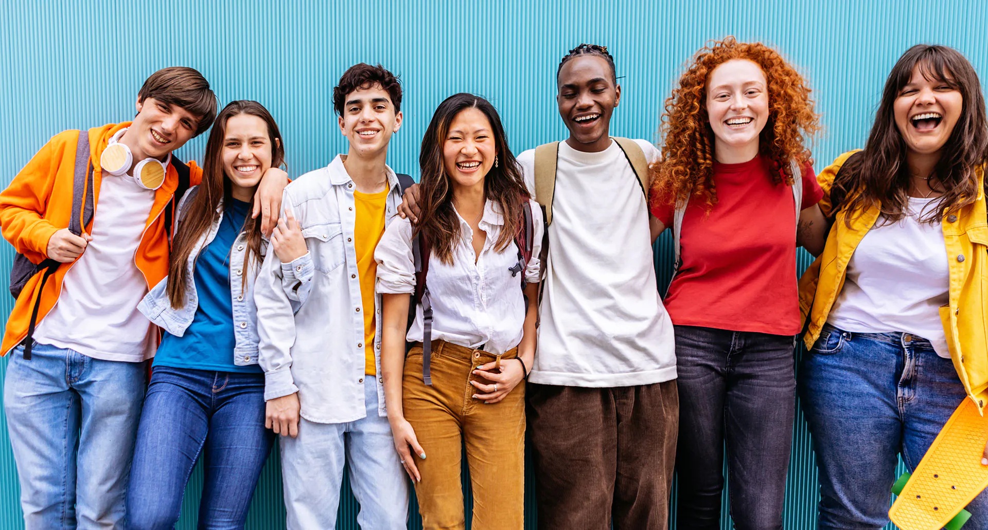 A group of seven young people stand shoulder to shoulder in front of a turquoise wall. They are all smiling broadly and look happy; they are wearing colourful clothes and backpacks, and the atmosphere is friendly and cheerful.