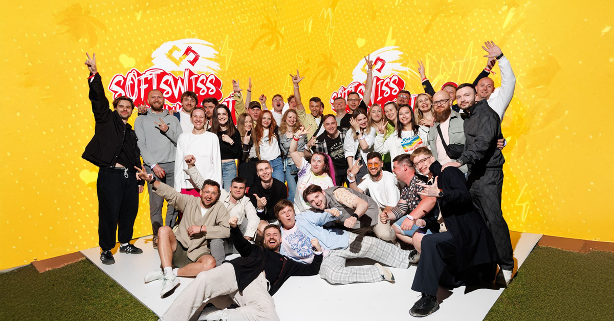 A large group of people pose in front of a yellow wall. They are smiling and happy.