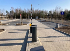 At the centre there is an entry barrier with a parking payment machine. The surface is made of paving stones, with clearly marked traffic lanes and parking spaces. On the right-hand side, a mandatory one-way traffic sign is visible. The car park is empty, with no vehicles parked.