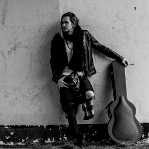 Black and white photo of the vocalist, who stands leaned against a wall. He holds a guitar case, which is also leaning against the wall.