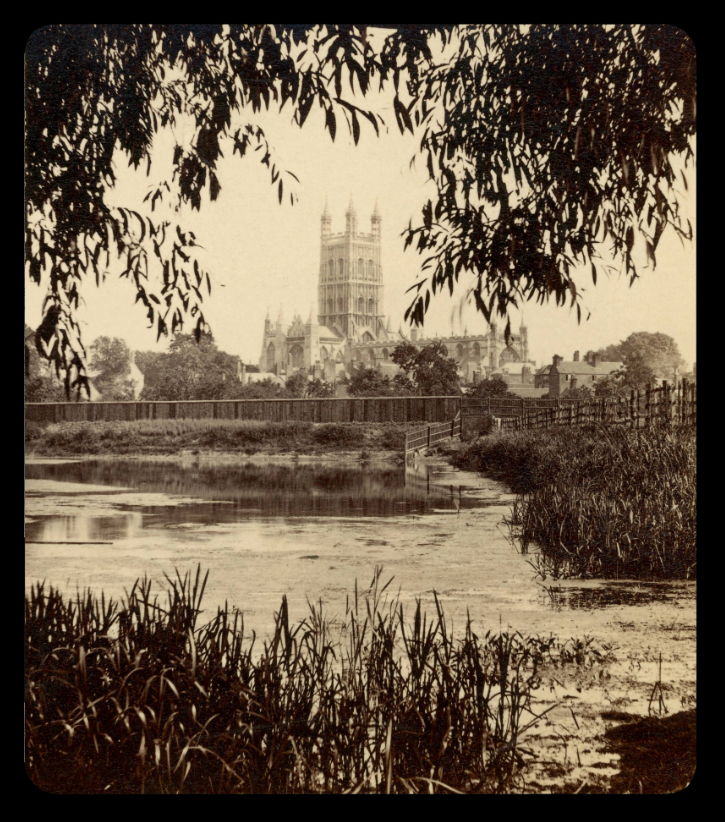 Sepia photograph of the Gloucester Cathedral, which is in the background. In the foreground a lake or a river with some water plants.