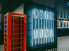 It's the interior of a cinema with a modern retro style. A large neon sign reading "KINO MUZA" glows on the wall. Next to it stands a red British-style phone booth. Illuminated movie posters hang on the walls, and the floor features a black-and-white checkerboard pattern. Dark tones dominate the space, accented by bright neon lights, creating a moody, artistic atmosphere.