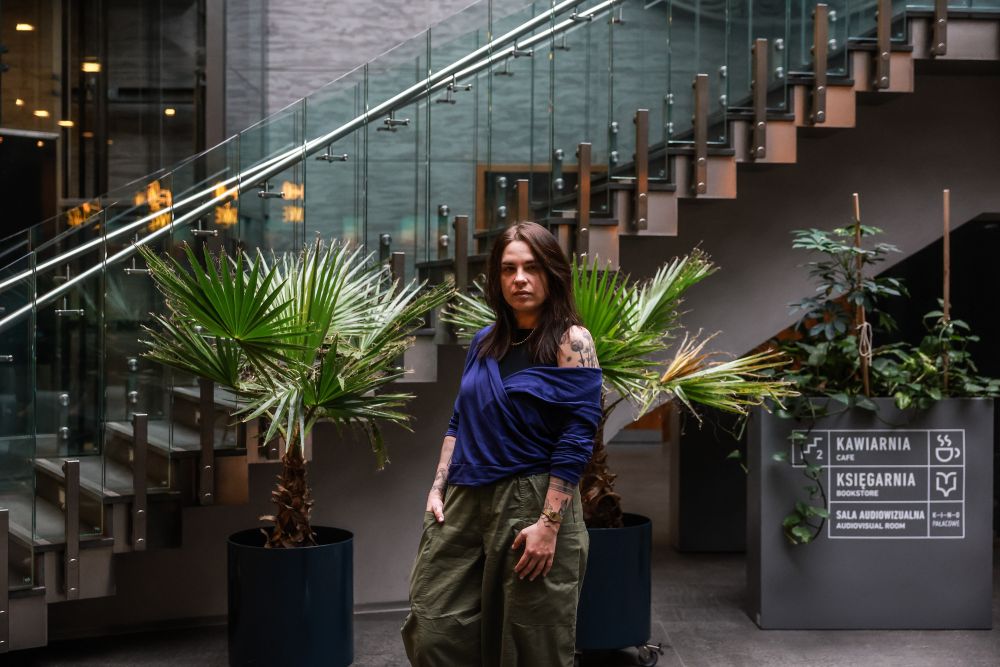 Aga Zano ma brązowe włosy do ramion, jest ubrana w niebieską bluzkę z opadającym jednym rękawem oraz luźne zielone spodnie. Siedzi na szarej kanapie, trzymając nogę na nodze.