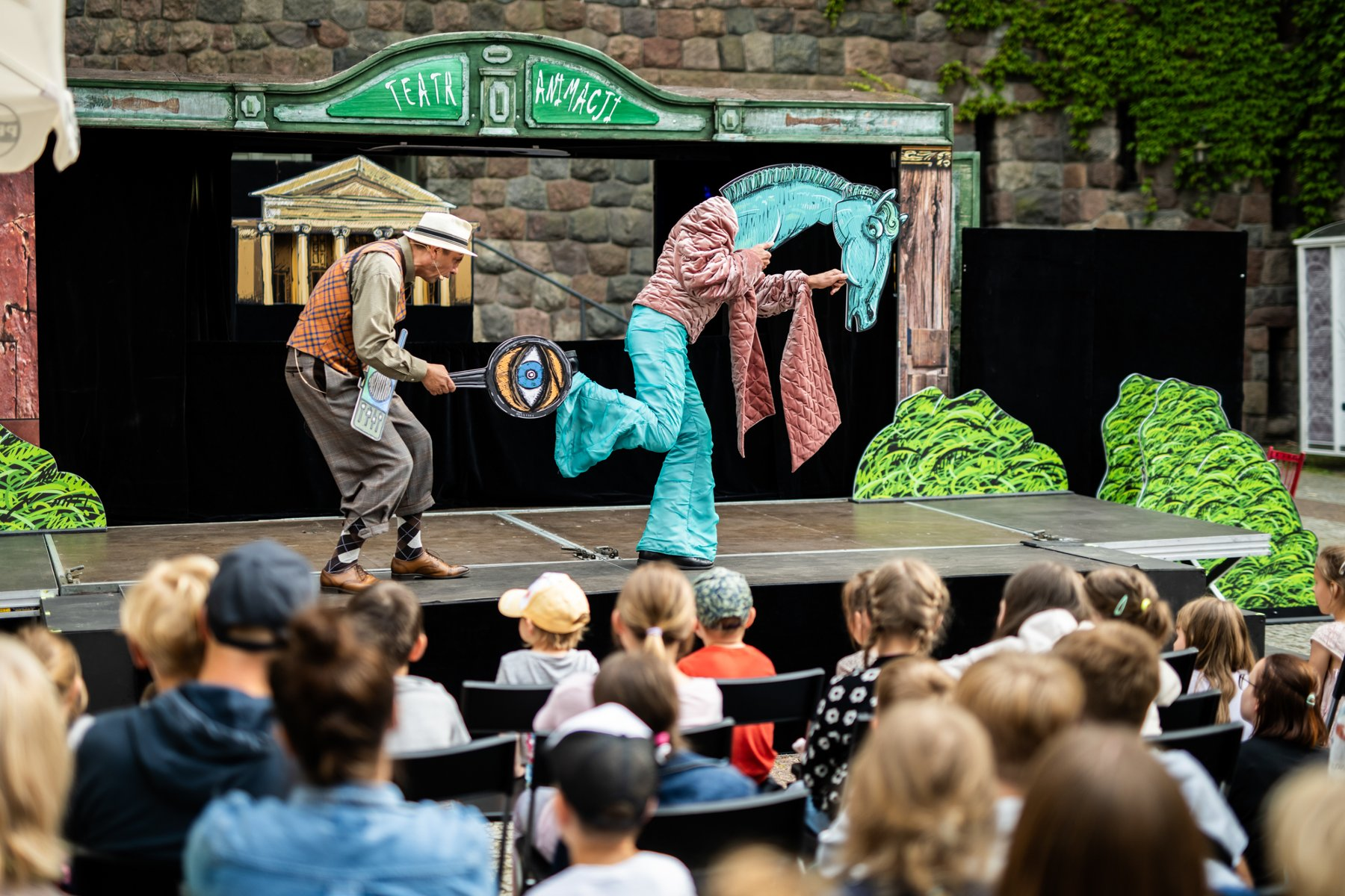 Aktorzy Teatru Animacji na scenie. Mężczyzna w kapeluszu trzyma patelnię z dużym okiem, a kobieta w kostiumie konia ze zwisającymi różowymi szalami pozuje. Na tle sceny dekoracje stylizowane na budynek z kolumnami i zielone pagórki. Na górze sceny napis "TEATR ANIMACJI". Przed sceną widać z tyłu głowy dzieci i dorosłych oglądających przedstawienie.