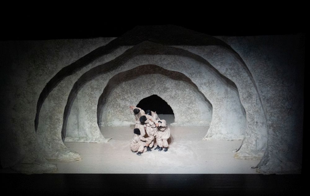 Artystka ma na sobie białą pelerynę, trzyma jej poły w dłoniach, oświetla ją mocne światło, a ona jakby wynurza się z ciemności.