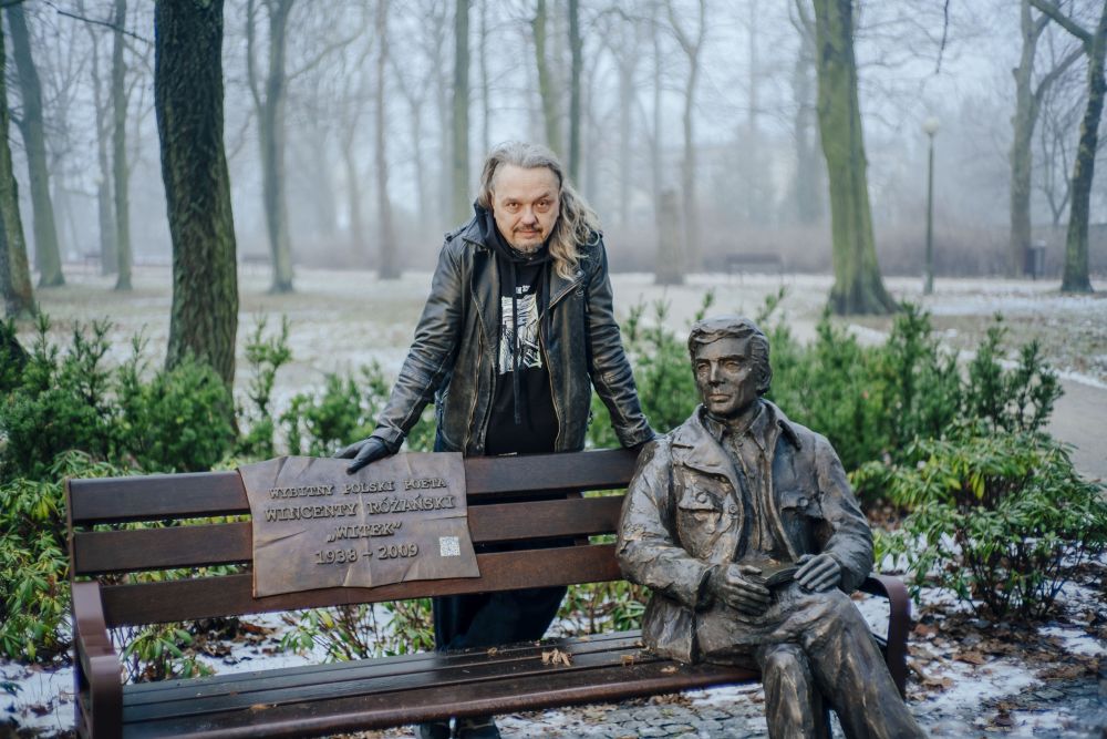 Autor ma na sobie czarną skórzaną kurtkę i motocyklowe rękawiczki, ma długie włosy do ramion.