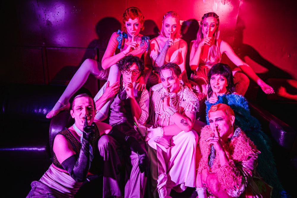 Barwnie ubrani aktorzy kładą palce na usta w geście oznaczającym ciszę.