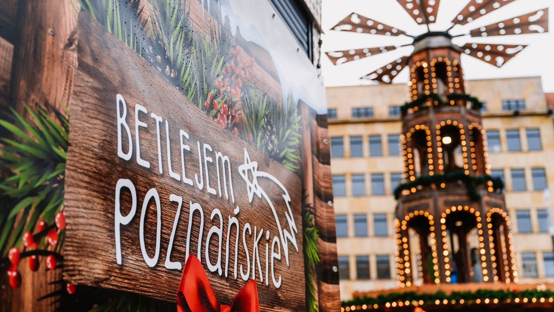 Po lewej napis na brązowej tabliczce "Betlejem Poznańskie". W tle karuzela i widok na budynek na placu Wolności.