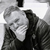 Mężczyzna siedzi w kurtce skórzanej i dłonią zakrywa usta.