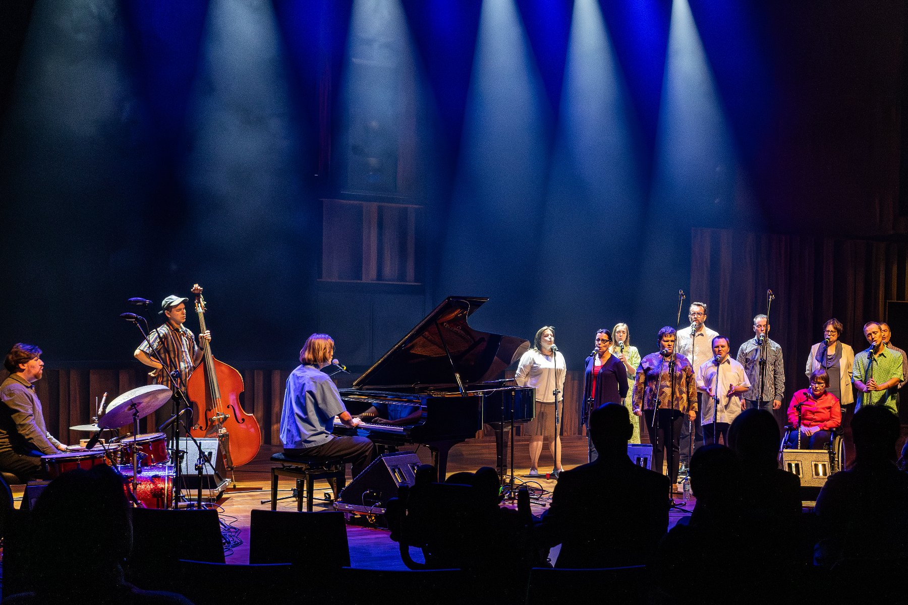 Chórzyści przy mikrofonach. Ubrani po cywilnemu, w różnym wieku. Kilka osób z niepełnosprawnością intelektualną. Po lewej stronie orskietra. Jest perkusja, kontrabas, fortepian.