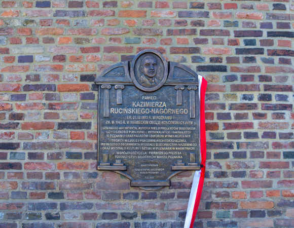 Ciemna, metalowa tablica pamiątkowa zamontowana na ceglanym murze. U góry tablicy znajduje się płaskorzeźba z portretem mężczyzny. Poniżej widnieje napis: "PAMIĘCI KAZIMIERZA RUCIŃSKIEGO-NAGÓRNEGO", a pod nim szczegółowy opis jego zasług jako architekta. Z prawej strony tablicy przewieszona jest biało-czerwona wstęga.