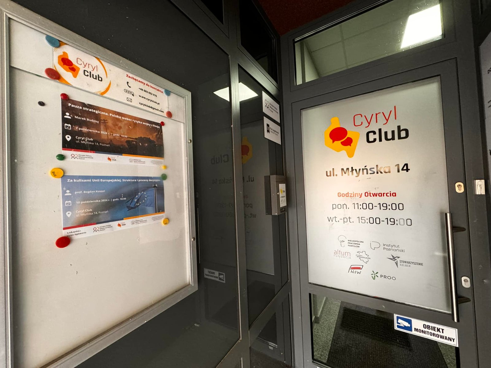 Zdjęcie przedstawia wejście do klubu o nazwie "Cyryl Club" w Poznaniu, Polska. Klub znajduje się na ulicy Młyńskiej, numer 14. Na drzwiach znajduje się tabliczka z nazwą i adresem klubu, a także godzinami otwarcia. Po lewej stronie od wejścia znajduje się tablica z plakatami reklamującymi nadchodzące wydarzenia, a także czerwone i niebieskie magnesy. Dodatkowo na drzwiach znajduje się naklejka informująca, że lokal jest monitorowany.