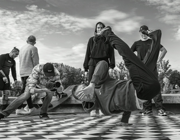 Czarno-białe zdjęcie przedstawiające grupę młodych mężczyzn na zewnątrz. Na pierwszym planie tancerz breakdance wykonuje akrobatyczny ruch, stojąc na dłoni na posadzce w czarno-białą kratę. Obok niego, inny mężczyzna klęczy, nagrywając jego występ kamerą. W tle stoi czterech innych obserwatorów.