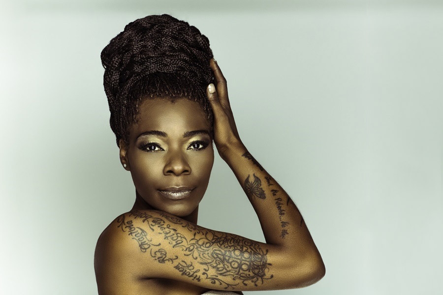 Czarnoskóra artystka patrzy w obiektyw, prawą dłoń przykłada do włosów. Jej ramię pokrywają tatuaże.