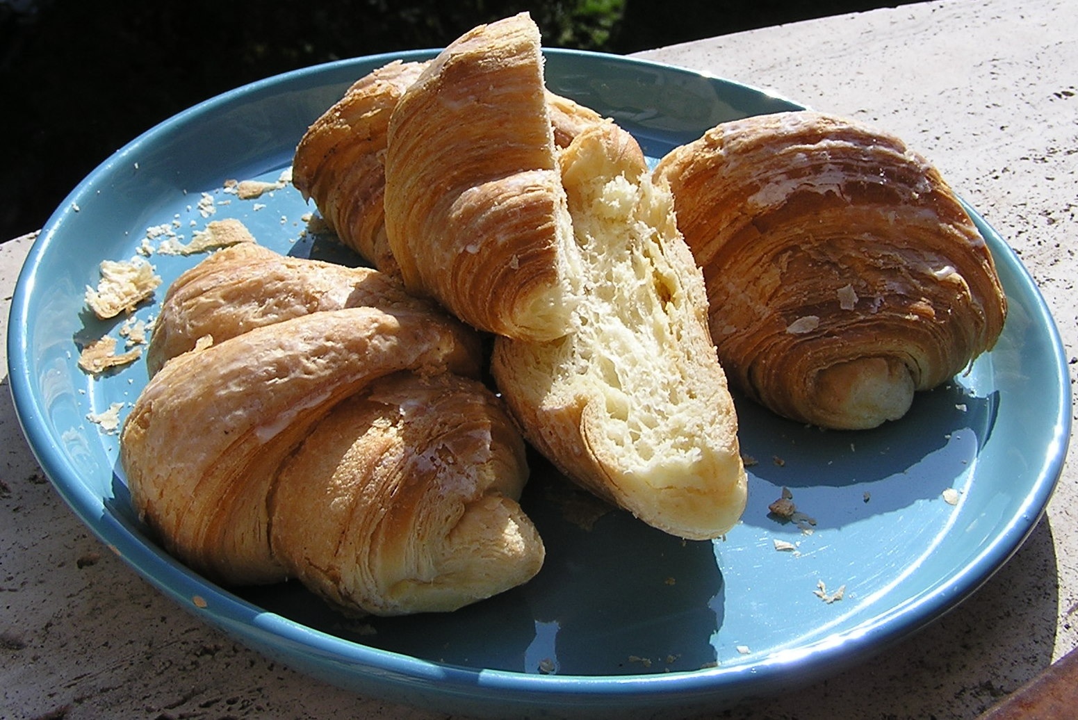 Cztery złociste, świeże rogaliki na turkusowym talerzu. Jeden z rogalików jest przekrojony na pół, ukazując jego jasny, puszysty środek. Wokół rogalików widać okruszki. Talerz stoi na kamiennym blacie w ostrym słońcu.