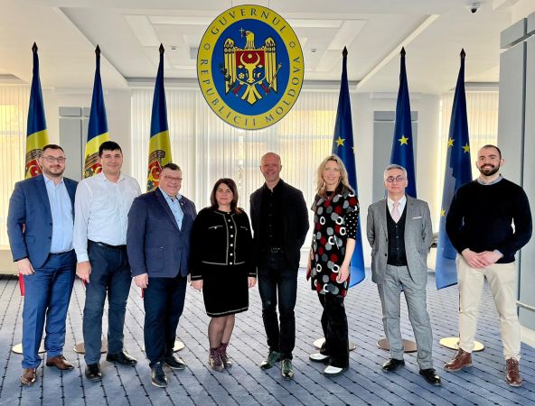 Delegacja ośmiu osób pozujących pod godłem Republiki Mołdawii, między flagami Mołdawii i Unii Europejskiej. Wszyscy ubrani elegancko, stoją w świetle dziennym.