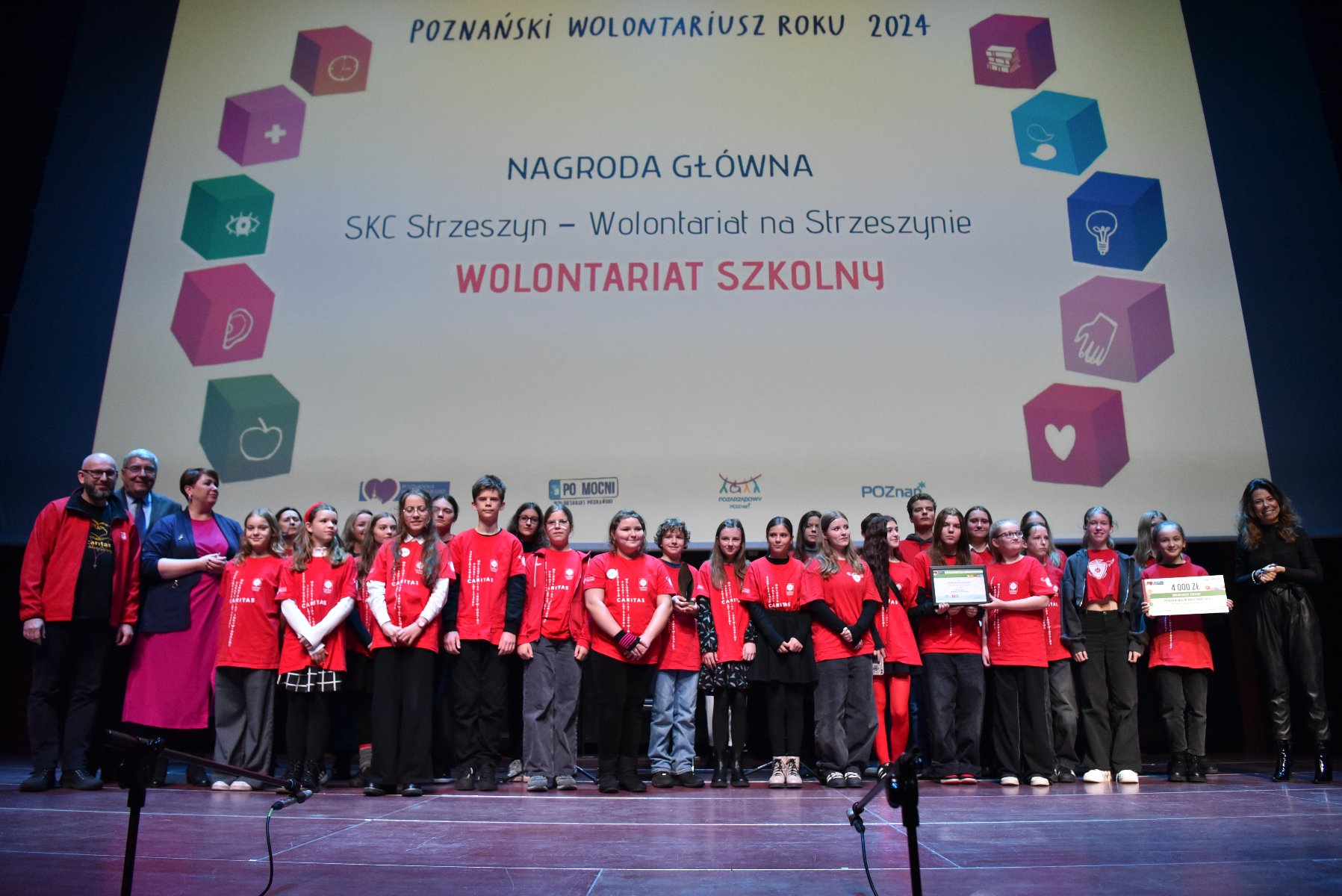 Duża grupa dzieci i młodzieży w czerwonych koszulkach stoi na scenie obok dorosłych podczas gali finałowej konkursu "Poznański Wolontariusz Roku 2024". Za nimi wisi ekran projekcyjny z widocznym napisem: "NAGRODA GŁÓWNA SKC Strzeszyn - Wolontariat na Strzeszynie WOLONTARIAT SZKOLNY" oraz dekoracyjnym łukiem złożonym z kolorowych kostek z symbolami (serce, jabłko, ucho, krzyż, mózg, żarówka). Dzieci trzymają dyplom i duży czek z kwotą 9 000 zł.