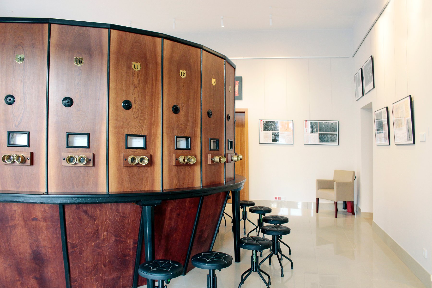 Duża, półokrągła, drewniana szafa z licznymi przegródkami i lunetami. Na górze każdej przegródki widnieje złota cyfra. Przed szafą stoją czarne, okrągłe stołki, a w tle widać jasną salę wystawową z obrazami na ścianach i beżowym fotelem.