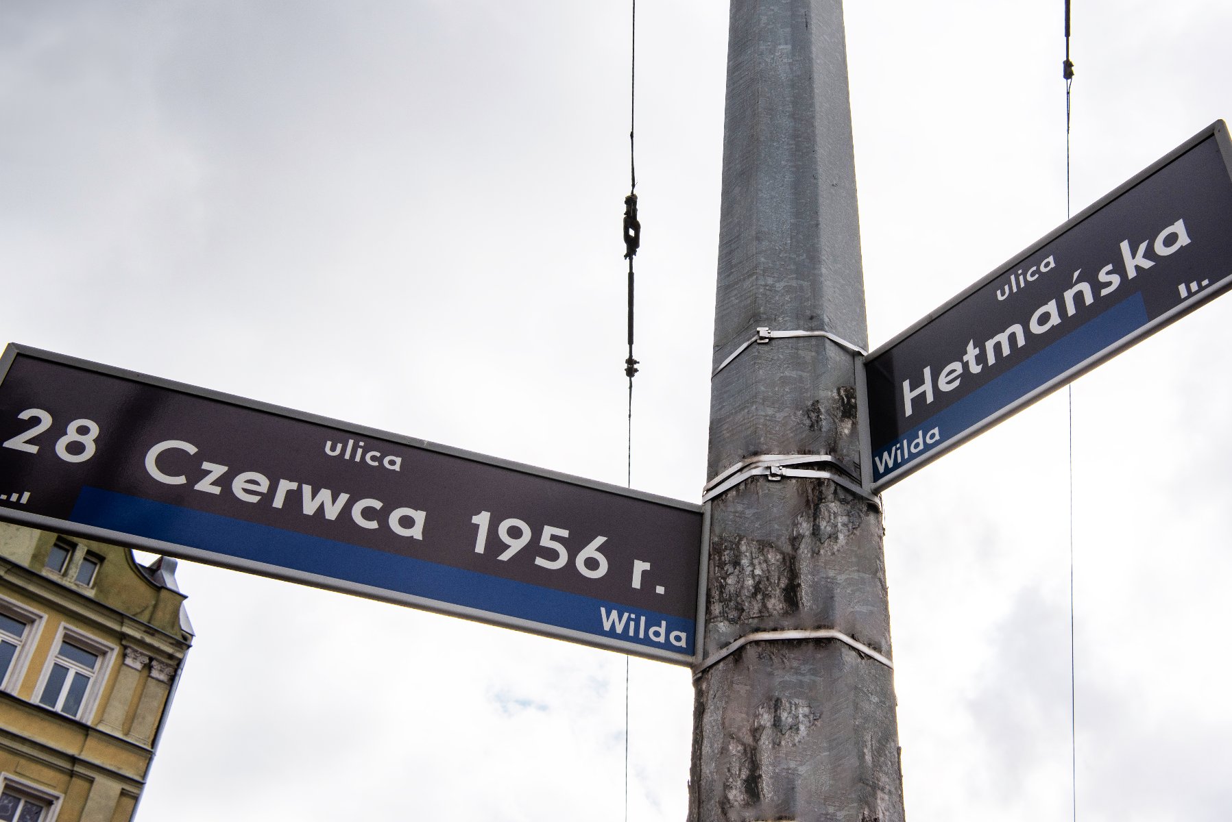 Dwa czarne znaki drogowe z białymi napisami. Na jednym widnieje napis "ulica Hetmańska Wilda", a na drugim "ulica 28 Czerwca 1956 r. Wilda". Znaki przymocowane są do szarego słupa, w tle widać niebo.