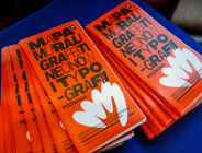 Dwa stosy pomarańczowych ulotek lub broszur rozłożone na niebieskim podłożu. Na okładkach widnieje duży, czarny napis w języku polskim: »MAPA MURALI, GRAFFITI, NEONÓW I TYPOGRAFII POZNANIA«. Poniżej znajduje się biała grafika przypominająca dłoń lub kwiat oraz mniejszy napis w języku angielskim.