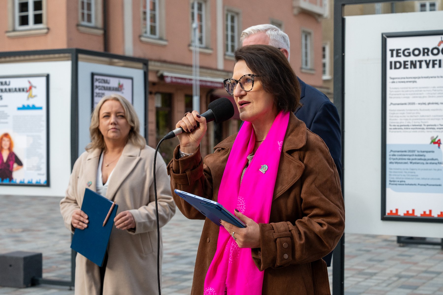 Dwie kobiety na tle wystawy na placu jedna z kobiet na pierwszym planie trzyma w ręku mikrofon za nią mężczyzna słabo widoczny na zdjęciu