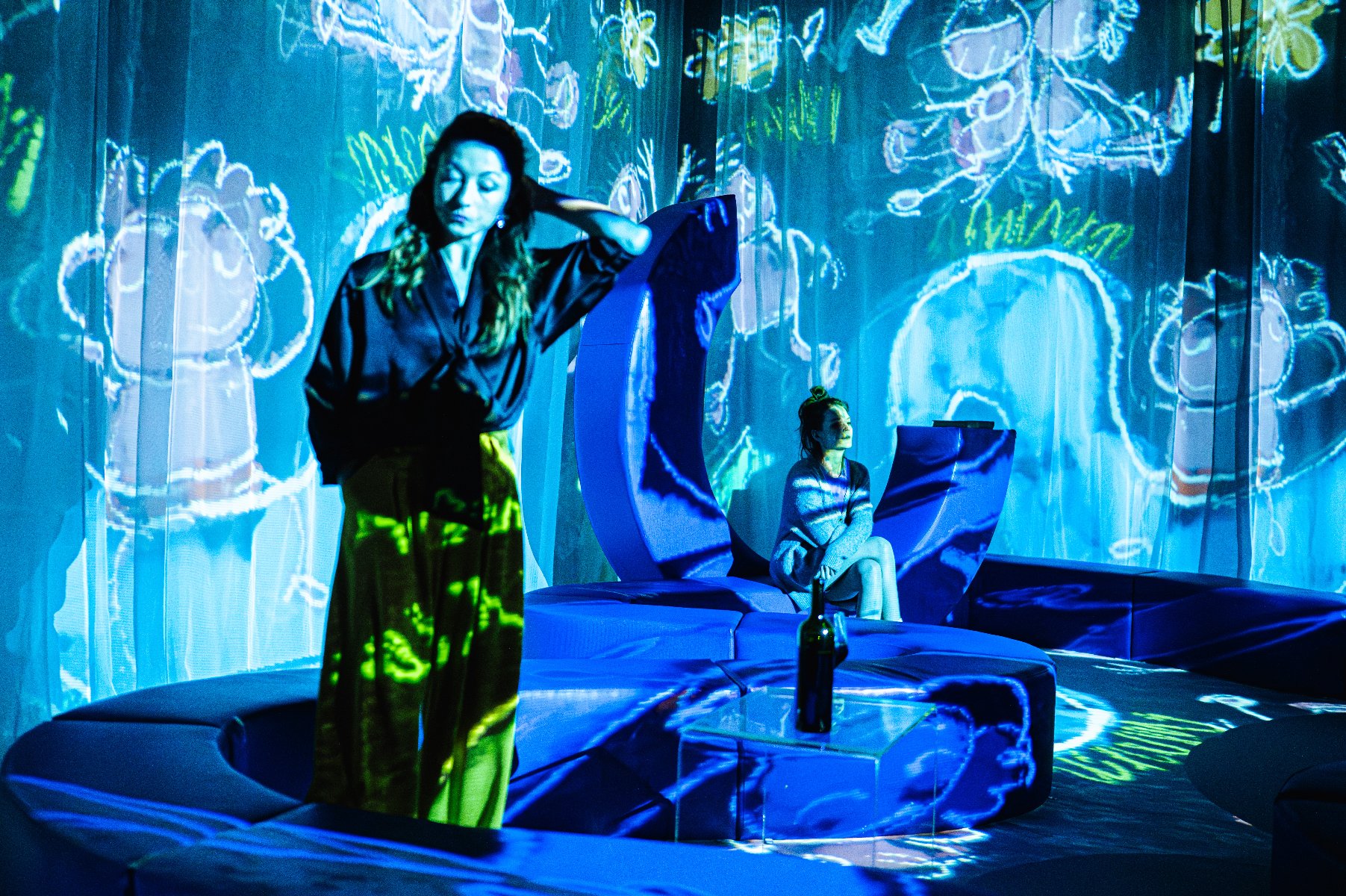 Dwie młode kobiety w strojach domowych na tle wideo z rysunkami dziecią narysowanych kredą. na stoliku butelka wina.