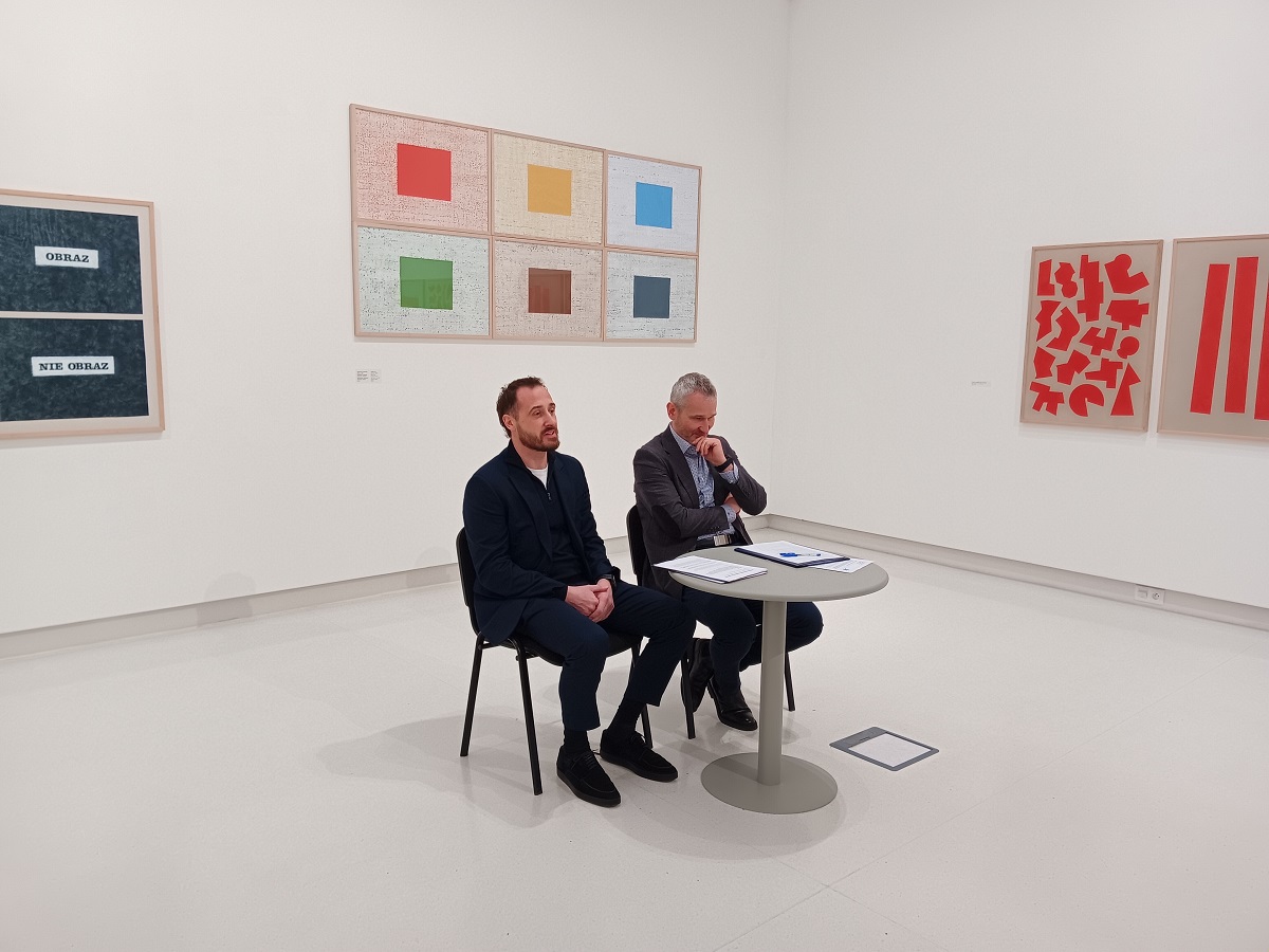 dwóch mężczyzn siedzi przy stoliku w białej sali, na jej ścianach wiszą obrazy przedstawiające sztukę nowoczesną