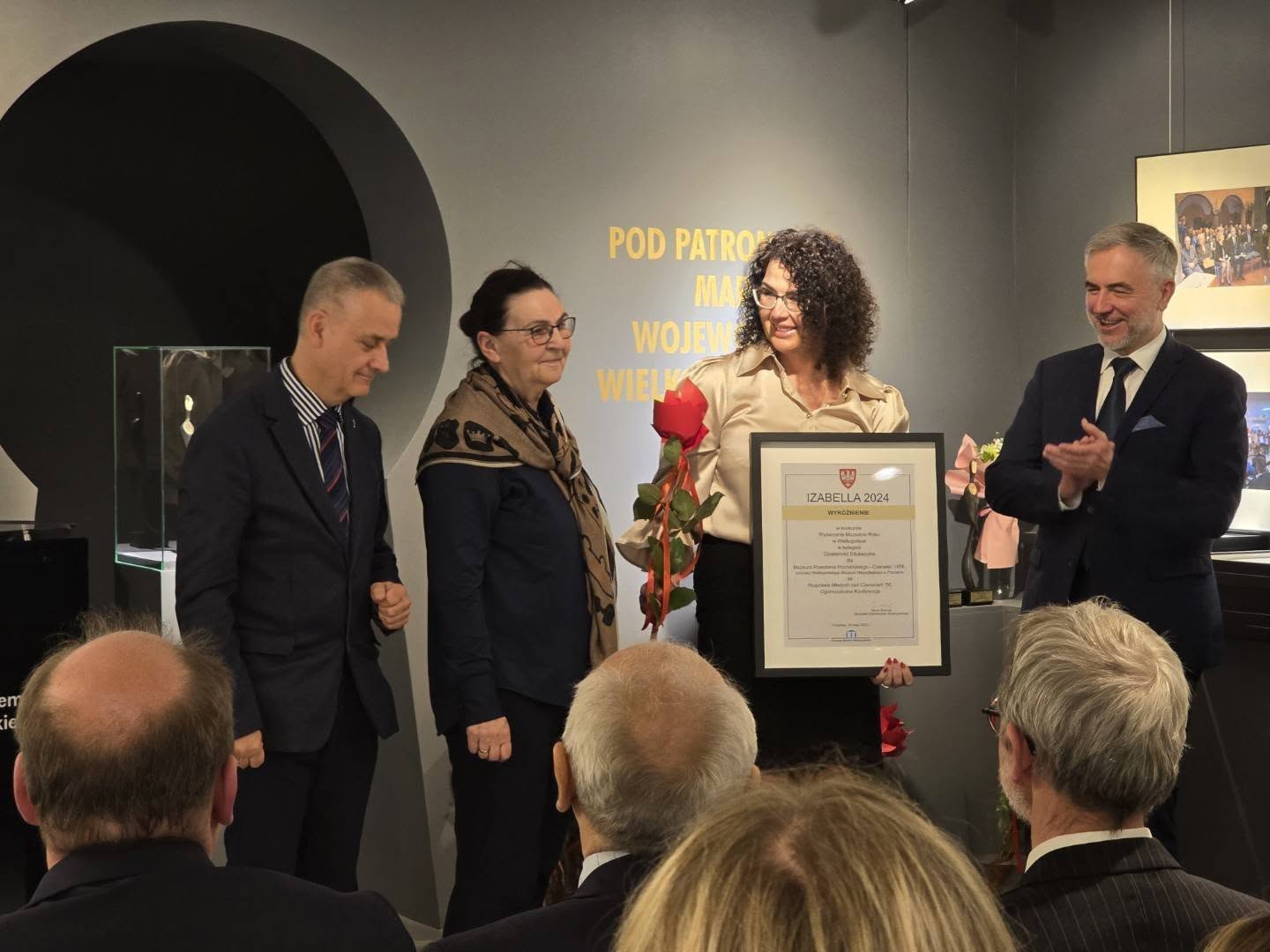 Dwóch mężczyzn ubranych w garnitury i dwie kobiety ubrane na galowo. Jedna z kobiet trzyma tabliczkę z nagrodą oraz czerwoną różę. Mężczyzna po prawej bije brawo. Kobieta i mężczyzna po lewej pozują do zdjęcia.