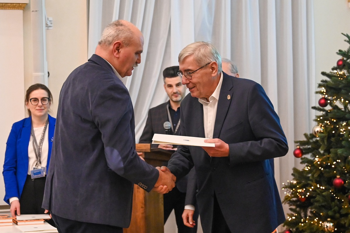 dwóch mężczyzn w garniturach jeden przekazuje drugiemu gratulacje i wyróżnienie - więcej w treści artykułu