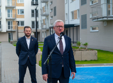 Dwóch mężczyzn w granatowych garniturach stoi przed nowoczesnym budynkiem mieszkalnym. Starszy mężczyzna w okularach przemawia do mikrofonu na stojaku. Na pierwszym planie widoczny fragment niebieskiej nawierzchni parkingu.