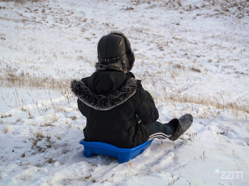 Dziecko ubrane w czarną, puchową kurtkę siedzi na niebieskim siedzisku i zjeżdża z górki. Na ziemi leży biały śnieg.
