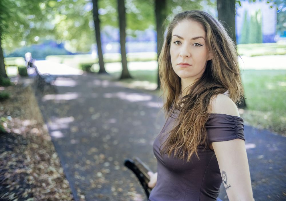 Dziewczyna oparta o drzewo, otaczają ją zielone liście, za nią park w letni, słoneczny dzień.