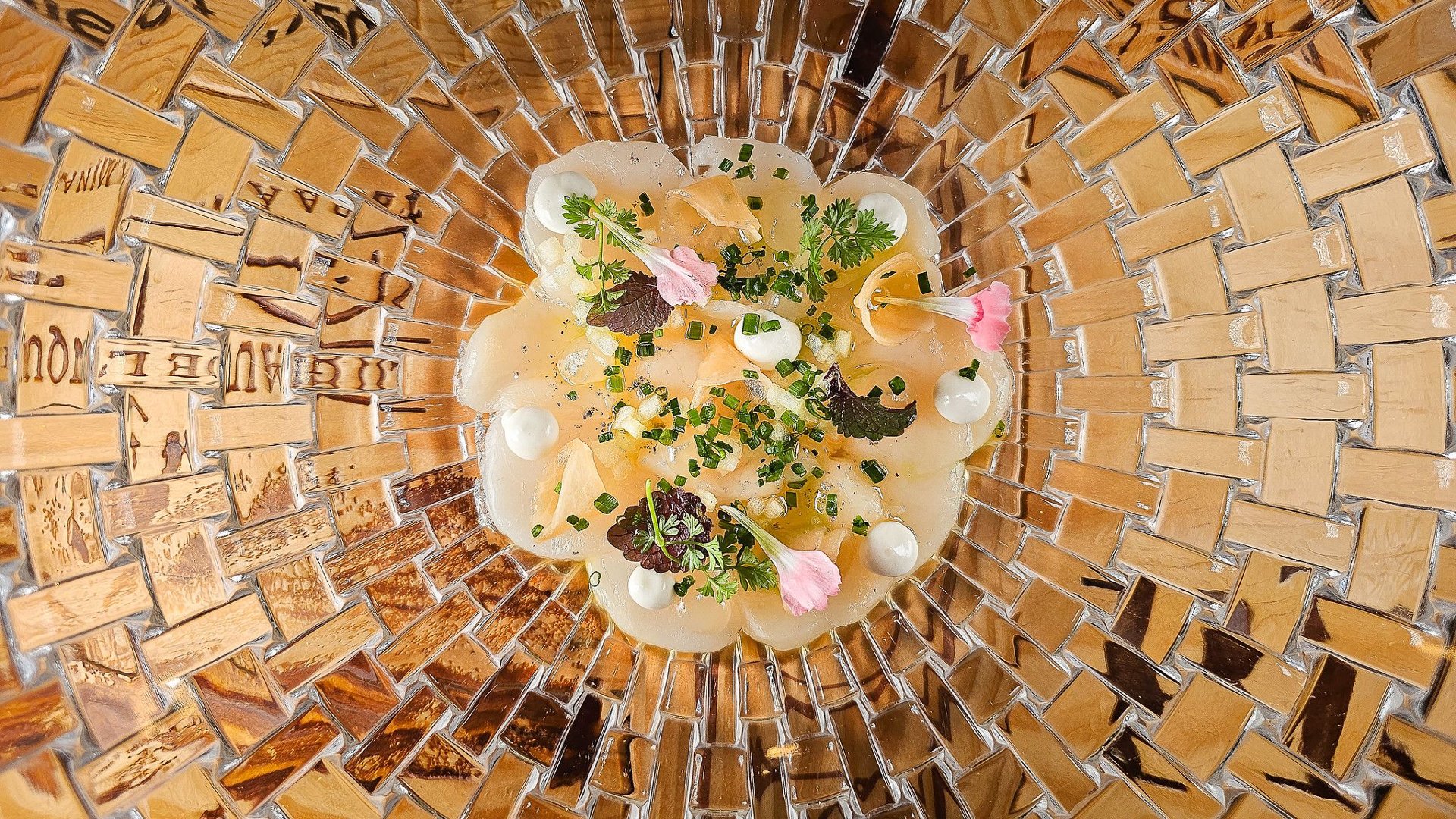 Elegancko podane danie w stylu fine dining, przedstawiające cienko krojone plastry surowej ryby (najprawdopodobniej carpaccio z przegrzebków lub ryby), udekorowane kremowymi kroplami sosu, drobno siekanym szczypiorkiem, listkami ziół i jadalnymi kwiatami. Całość podana w misie o fantazyjnym, mozaikowym wzorze z przezroczystego szkła przypominającego drewnianą strukturę.