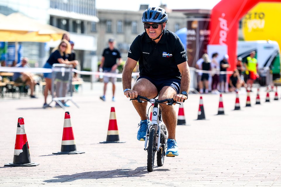 Enea IRONMAN 70.3 Poznań, fot. Przemek Świderski/ Sport Evolution - na małym rowerze jedzie mężczyzna w kasku i czarnym stroju. W tle widać pachołki oraz obsługę zawodów