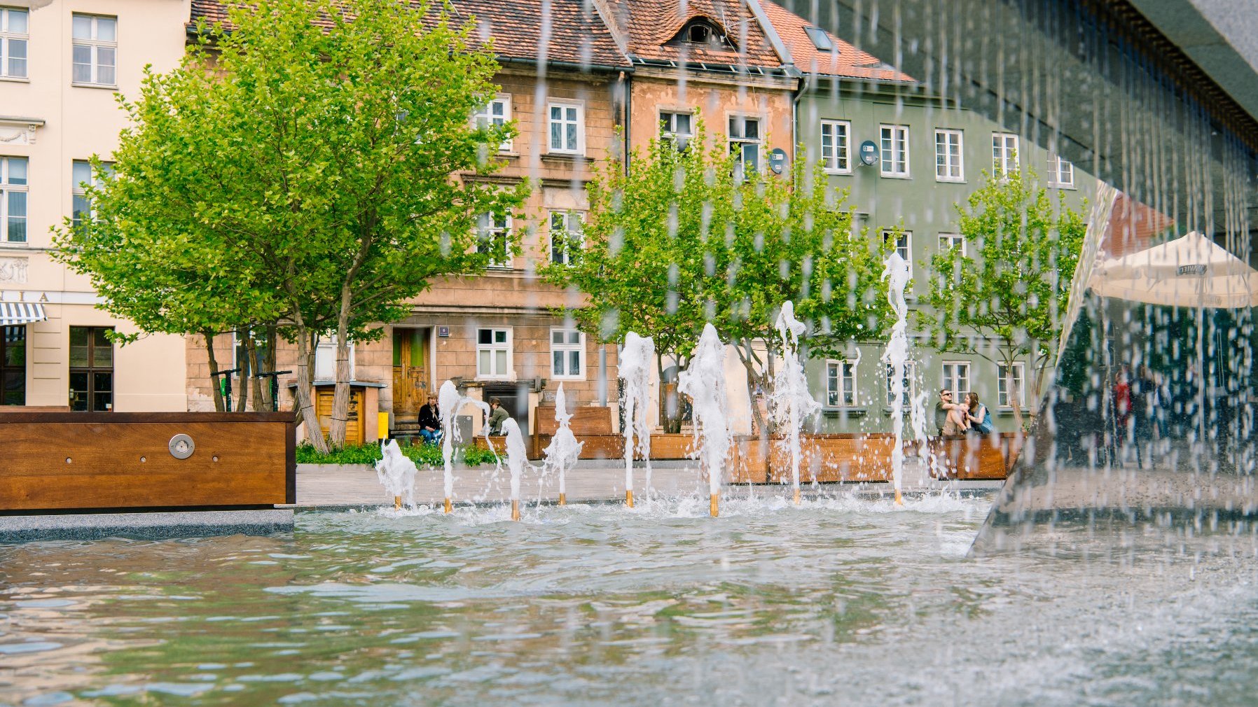 Fontanna wodna przy placu Kolegiackim. Z wodospadów spływa woda do zbiornika. W tle zielone drzewa i kolorowe kamienice.