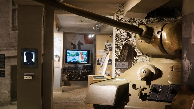 Ekspozycja w Muzeum: po lewej na scianie monitor z wyswietlona postacią, w dłebi duży monitor, po prawej czołg i duże zdjęcia na scianie postaci.