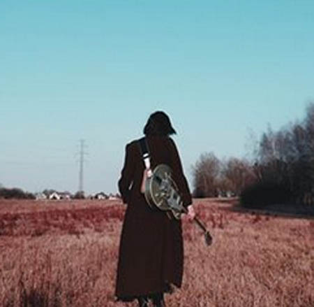 Foto. Kobieta z gitarą idzie przez pole.