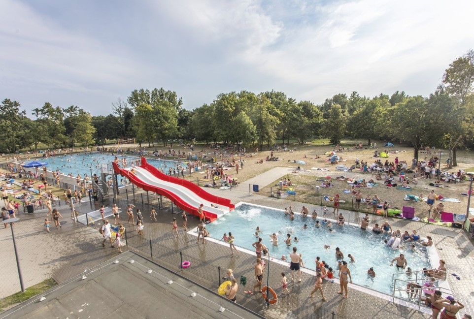 Fotografia przedstawia baseny w parku Kasprowicza ujęte na zdjęciu w oddali, widać również oddalone sylwetki ludzi w kostiumach kąpielowych korzystających z kapieliska