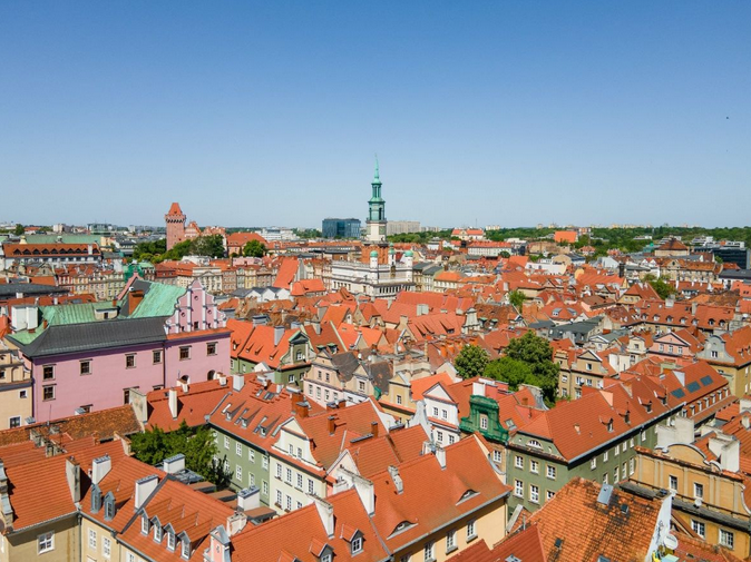 Fragment panoramy Starego Miasta w Poznaniu, widoczne dachy kamienic z czerwoną dachówką, w tle widoczna wieża ratusza i wieża zamku Przemysła, na miastem błękitne niebo, słoneczna pogoda