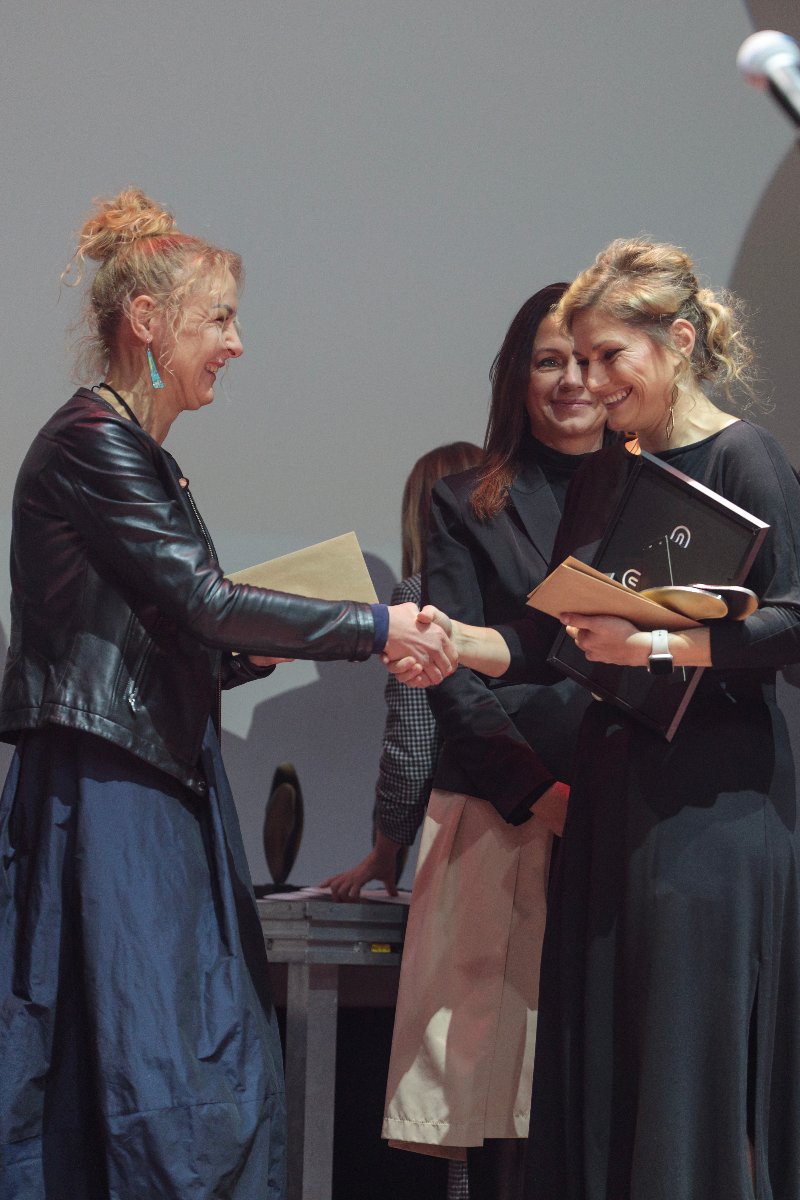 Na zdjęciu znajdują się 3 kobiety. Jedna z nich odbiera nagrodę, dwie pozostałe jej gratulują, jedna z gratulujących ściska rękę laureatce.