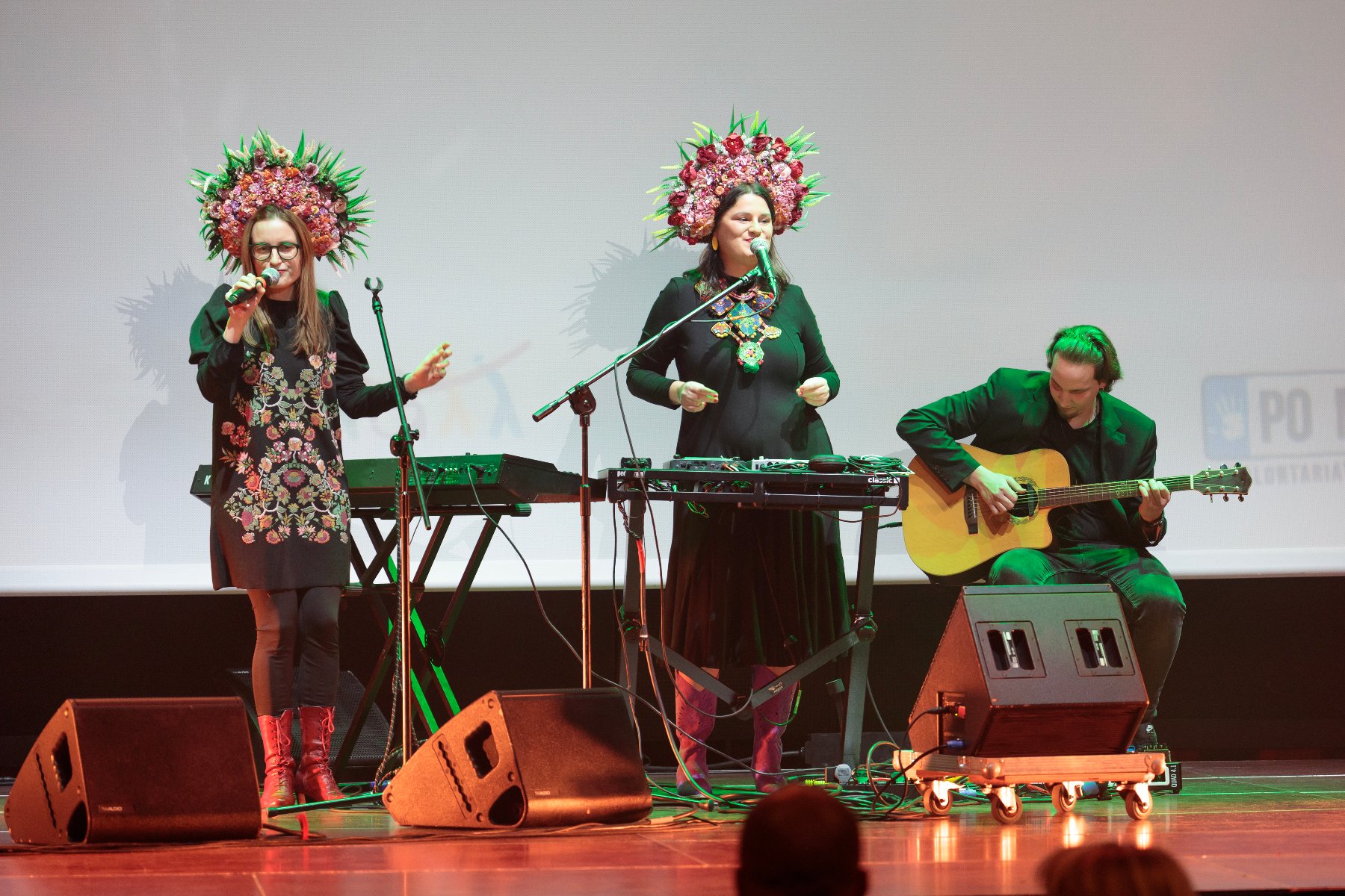 Na zdjęciu znajdują się dwie kobiety na scenie śpiewające do mikrofonów. Obie mają na głowach słowiańskie wieńce. Obok nich siedzi mężczyzna grający na gitarze.