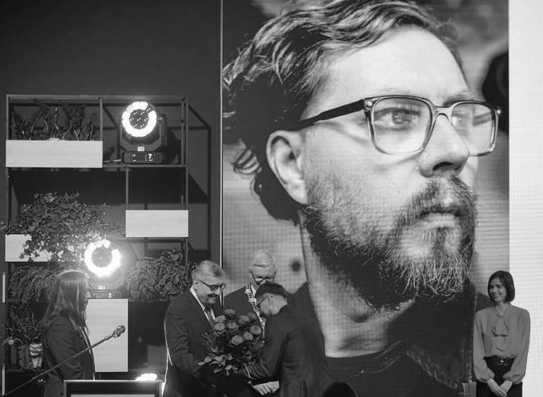 Gala wręczenie nagrody. Urzędnicy i bohater. W tle jego duże zdjęcie.