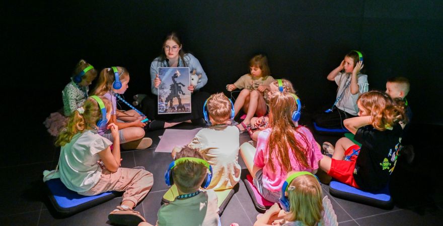 Grupa dzieci wraz z animatorką siedzą w kręgu i słuchają opowieści o koziołkach.