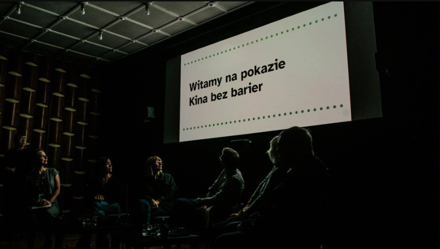 Grupa ludzi siedzi w ciemnej sali, zwróceni w stronę dużego ekranu. Na ekranie wyświetla się tekst po polsku: "Witamy na pokazie Kina bez barier". Sala wygląda na kinową lub prezentacyjną, z wzorzystą ścianą i oświetleniem sufitowym.
